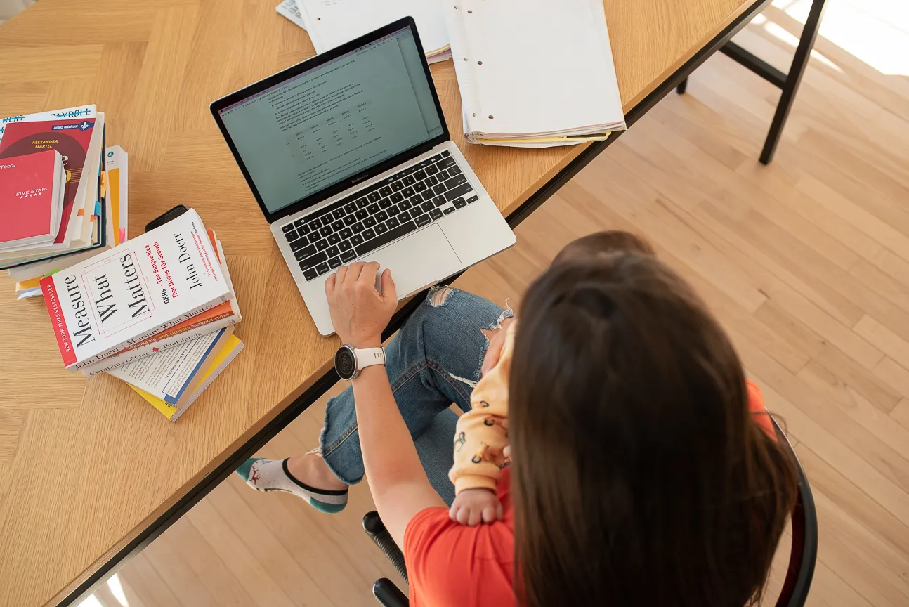Photo d'Hélène-Sarah Becotte à son bureau de travail avec son bébé dans les bras et des livres de mathématiques